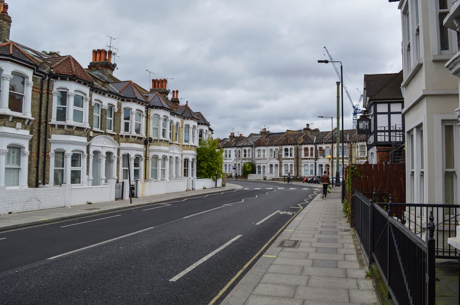 A quiet residential street in Roehampton shows a row of Victorian-style terraced houses with white facades, bay windows, and red-tiled roofs. The street is paved with asphalt and marked with white lin