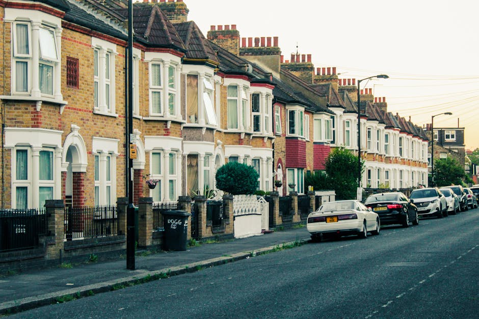 A quiet residential street in Roehampton shows a row of Victorian-style terraced houses with white facades, bay windows, and red-tiled roofs. The street is paved with asphalt and marked with white lin